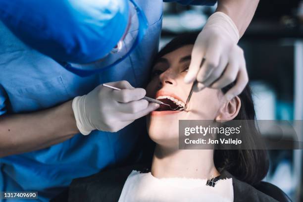 un dentista con bata azul, guantes de látex, gafas transparentes y mascarilla limpia los dientes de un paciente con una herramienta metálica. - root canal procedure stock-fotos und bilder