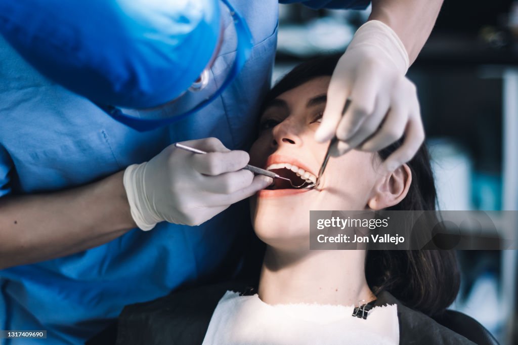 Un dentista con bata azul, guantes de látex, gafas transparentes y mascarilla limpia los dientes de un paciente con una herramienta metálica.