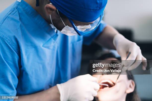 un dentista con bata azul, gafas de plástico transparentes y mascarilla utiliza dos herramientas metálicas para limpiar los dientes de una paciente. enfoque selectivo. - doctor paciente stockfoto's en -beelden