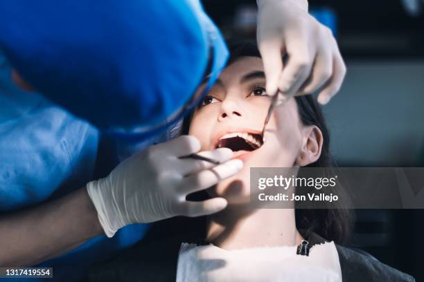 un dentista con guantes de látex, gafas transparentes y mascarilla abre la boca de una paciente con una herramienta metálica para observar sus dientes. - mascarilla stockfoto's en -beelden