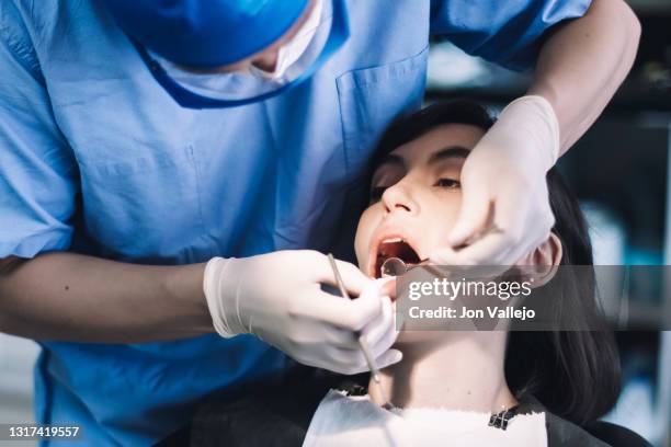 un dentista con guantes de látex, gafas transparentes y mascarilla observa los dientes de un paciente con un pequeño espejo metálico. - mascarilla stock pictures, royalty-free photos & images