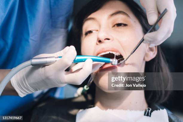 una paciente está con la boca abierta y un tubo de plástico succionando en su boca. el dentista sostiene un pequeño espejo metálico y una herramienta eléctrica en la mano. el dentista está a punto de introducir la herramienta eléctrica en la boca de - paciente stock pictures, royalty-free photos & images