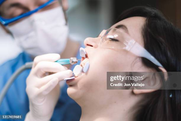 el odontólogo con gafas antisalpicaduras, mascarilla y bata azul está tratando la boca de una mujer joven con gafas antisalpicaduras de plástico y un separador dental. acercamiento selectivo a la paciente. - paciente stock pictures, royalty-free photos & images