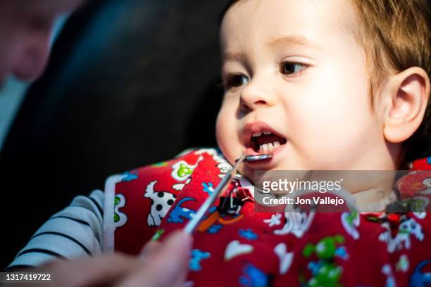 un niño de menos de dos años en la consulta del dentista, con una bata roja, mira al dentista mientras éste le baja el labio inferior con un pequeño espejo. enfoque selectivo. - 35 39 años stock pictures, royalty-free photos & images
