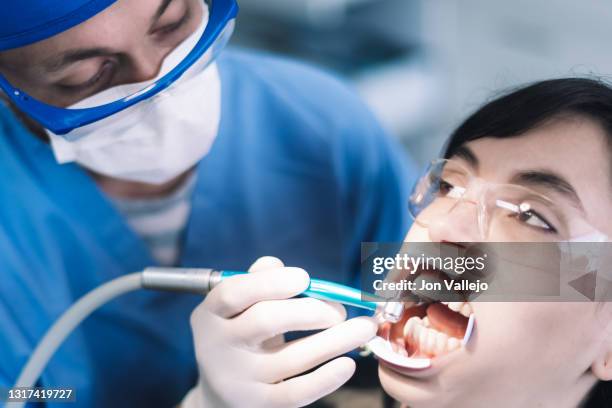 el dentista con sus gafas antisalpicaduras, mascarilla y bata azul está limpiando con una herramienta eléctrica los dientes de una joven que lleva gafas antisalpicaduras de plástico y un separador dental. acercamiento selectivo a la paciente. - mascarilla stock pictures, royalty-free photos & images