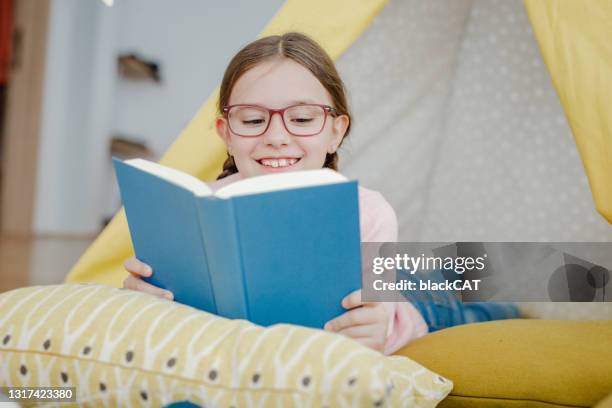 kleines mädchen liest ein buch unter ihrem selbstgebauten zelt im wohnzimmer - indoor kids play area stock-fotos und bilder
