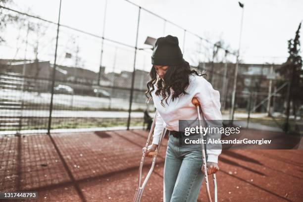mujer con muletas y sombrero de punto recuperando salud y forma en el campo deportivo - en muletas fotografías e imágenes de stock