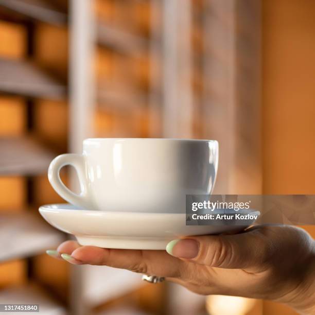 white porcelain or ceramic cup with saucer in female hand against window on blurred background. morning tea or coffee, care concept. close up shot. side view. soft focus - pires imagens e fotografias de stock