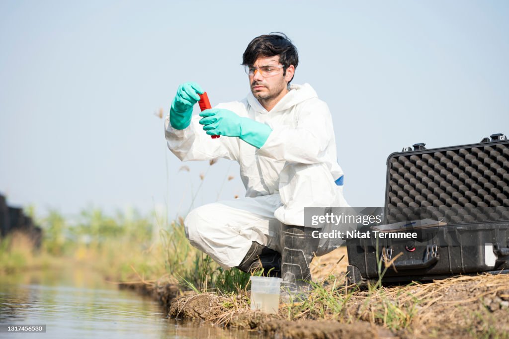 Male Scientist reading Ph value or examining toxic water from a water sample. He working outdoors and wearing protective workware coveralls. Water pollution and conservation and management of water concepts.