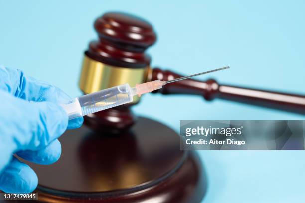 a person wearing surgical gloves holds a syringe. in the background a judge's gavel. - medical judge stock-fotos und bilder