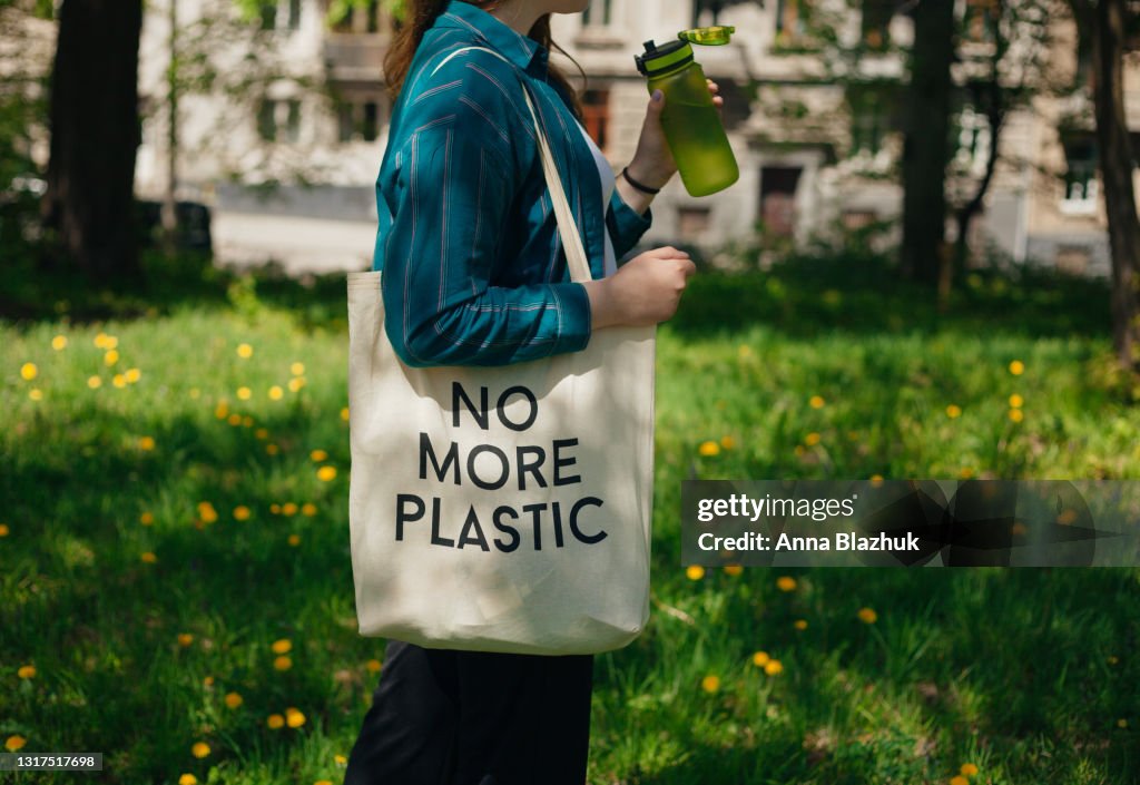 Woman holding reusable cotton zero waste bag with text No More Plastic. Outdoors portrait in sunny day. Eco friendly bags concept.