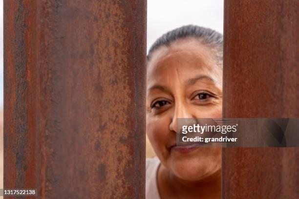 cheerful middle-aged adult mexican woman posing behind the international border barrier wall between the usa and mexico - international organisation for migration stock pictures, royalty-free photos & images
