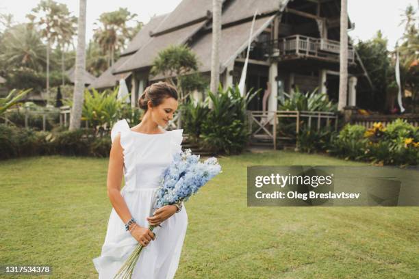 portrait of smiling woman in white linen dress with blue flower bouquet of delphinium - ranunculus wedding bouquet stock pictures, royalty-free photos & images