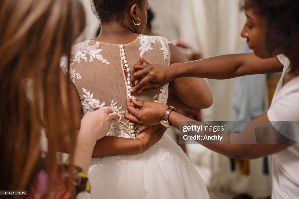 Woman trying on wedding dress with female friends having fun and helping her fit the dress