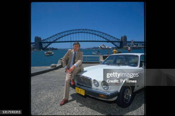 Television presenter Michael Parkinson sitting on the hood of a car with the Sydney Opera House and Harbour Bridge visible in the background, circa...