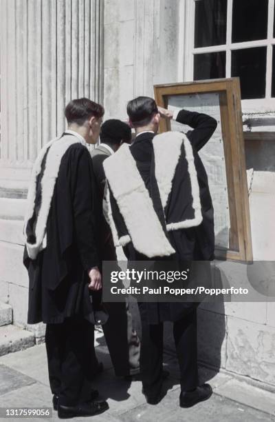 Wearing full academic dress comprising gown, hood and headdress, three graduates study degree exam results pinned to a board outside Senate House on...
