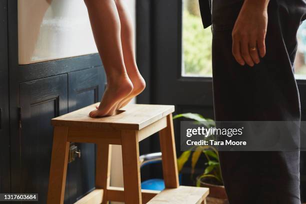 close up of legs and feet of a young mixed race girl, on tiptoes at kitchen sink on a low wooden stool. partial view of father behind her. - sgabello foto e immagini stock