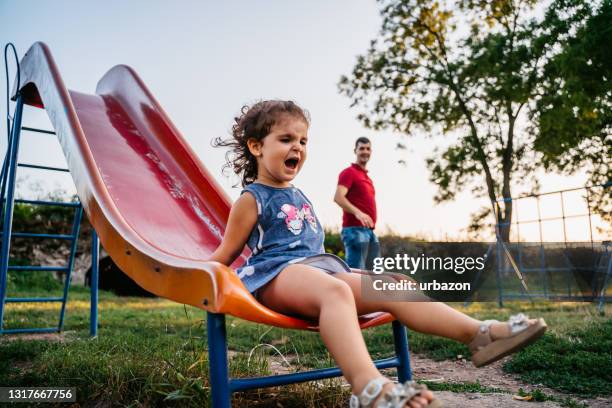 father watching out of little girl playing on playground slide - slide play equipment stock pictures, royalty-free photos & images