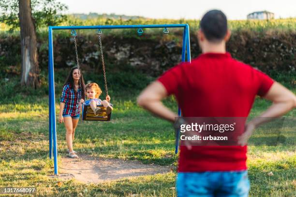 mother and daughter playing on swing and father watching them - playground equipment happy parent stock pictures, royalty-free photos & images