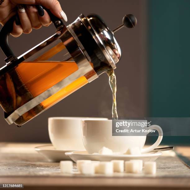 tea time. woman pouring hot golden drink from glass teapot or french press into cup. tea ceremony concept. side view - coffee plunger stock pictures, royalty-free photos & images