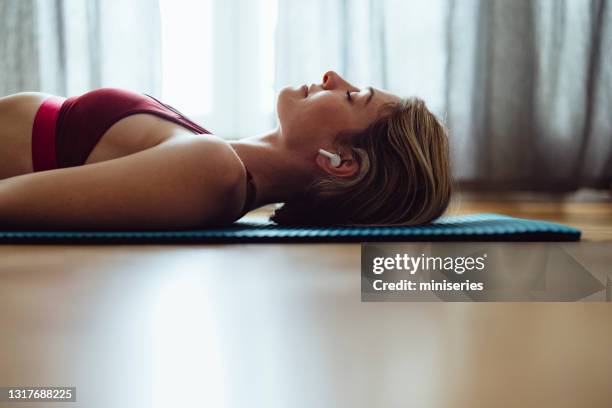 donna che medita sdraiata sul tappetino da esercizio indossando auricolari wireless - blonde yoga foto e immagini stock