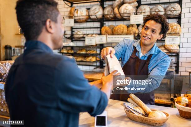 male staff giving takeaway food parcel to customer in a bakery - man offering bread stock pictures, royalty-free photos & images