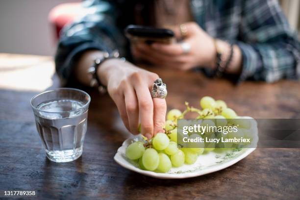 young woman with a unique style, using her smartphone and eating grapes - juicy lucy stock pictures, royalty-free photos & images