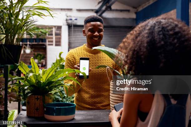 shot of a young man using a smartphone to pay for plants at a garden centre - digital payment imagens e fotografias de stock