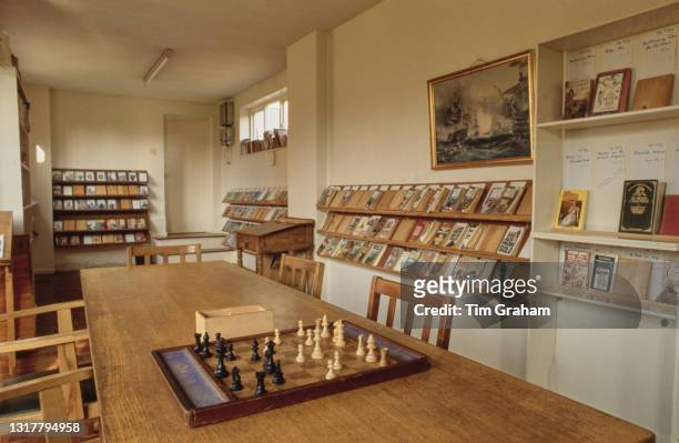 Framed painting hangs on the wall above the display shelves lined with books beyond a long table, on which is a chessboard, around which are chairs,...