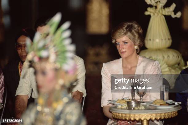 British Royals Charles, Prince of Wales, wearing a safari suit, partially obscured behind the headdress of a dancer, and Diana, Princess of Wales ,...