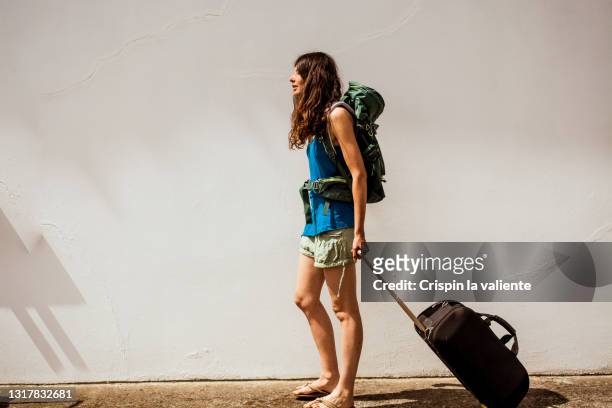 young traveler woman walking with backpack and hand suitcase on white background - long legs women stock-fotos und bilder