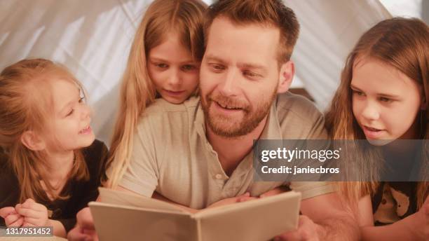 family reading time. father holding book and hugging his three daughters. - family with three children stock pictures, royalty-free photos & images