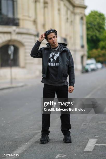 Alex Duarte wears black sunglasses, silver earrings, gold rings, a silver and rhinestones chain necklace, a black with white World print t-shirt with...