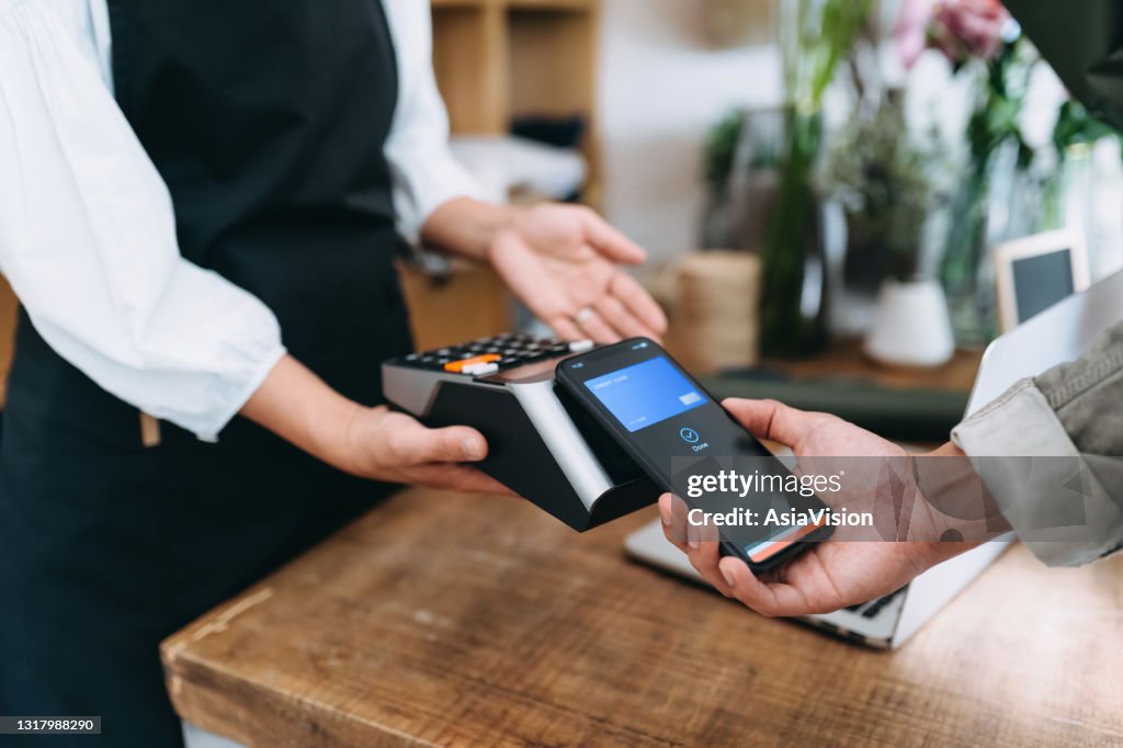 Närmd av unga asiatiska man som handlar i blomsteraffären. Han betalar med sin smartphone, skannar och betalar en räkning på en kortmaskin och gör en snabb och enkel kontaktlös betalning. NFC-teknik, tap and go-koncept