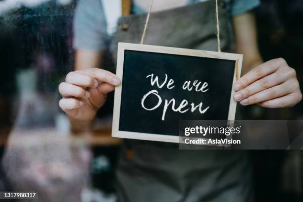 close up of young asian female small business owner in apron starting her business day. hanging an open sign on the door at the shop. business owner and everyday business practice concept - reopening sign stock pictures, royalty-free photos & images