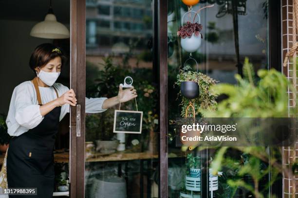 asian female florist, owner of small business flower shop wearing protective face mask, starting her business day. opening the door and hanging the open sign on her shop. new normal of everyday business practice - open practice stock pictures, royalty-free photos & images