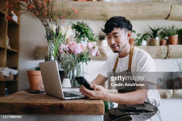 jonge aziatische mannelijke bloemist, eigenaar van kleine bedrijfsbloemenwinkel, die smartphone met behulp van terwijl het werken aan laptop tegen bloemen en installaties. voorraden controleren, bestellingen van klanten aannemen, producten online verkopen. - flower shop stockfoto's en -beelden