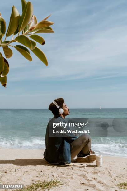 man with eyes closed listening music through headphones while sitting at beach - beach music stock pictures, royalty-free photos & images