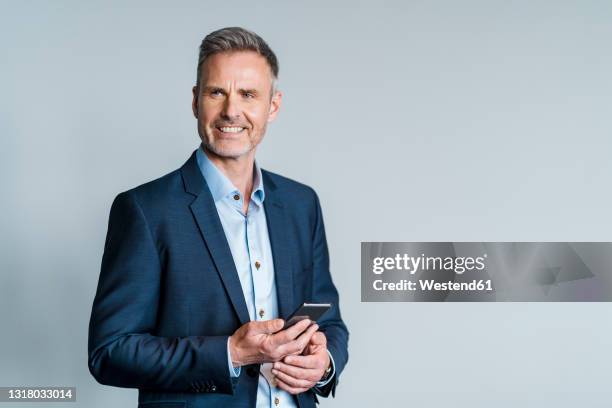 smiling male entrepreneur holding smart phone while standing by gray background - male cellphone stock-fotos und bilder