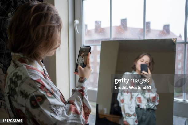 young woman photographing reflection in mirror through smart phone at home - ijdel stockfoto's en -beelden