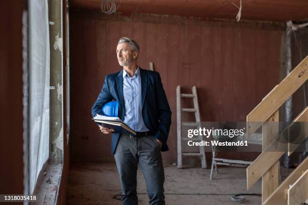 male architect with hand in pocket holding notebooks while looking through window at site - person in suit construction stock-fotos und bilder