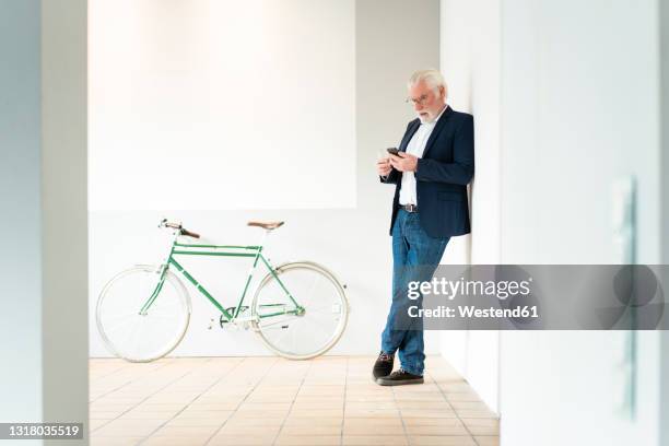 senior male entrepreneur using smart phone while leaning on wall at office - anlehnen stock-fotos und bilder