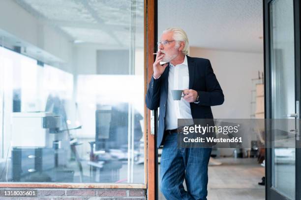 senior businessman smoking cigarette while leaning on glass wall at office - tobacco workers stockfoto's en -beelden