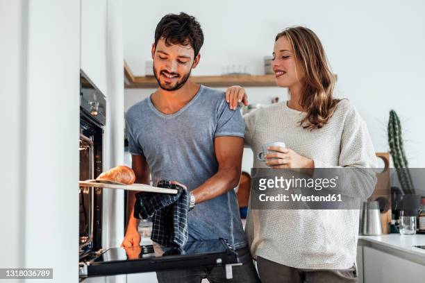 smiling woman looking at boyfriend removing baked croissant from oven in kitchen - man tray food holding stock pictures, royalty-free photos & images