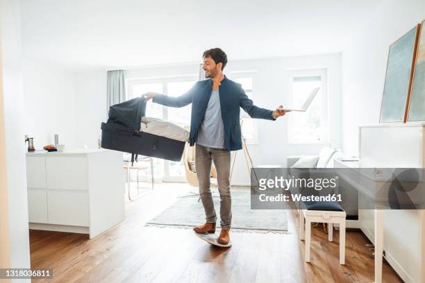 male freelancer carrying baby carrier and laptop on balance board in bright living room - multi tasking stock pictures, royalty-free photos & images