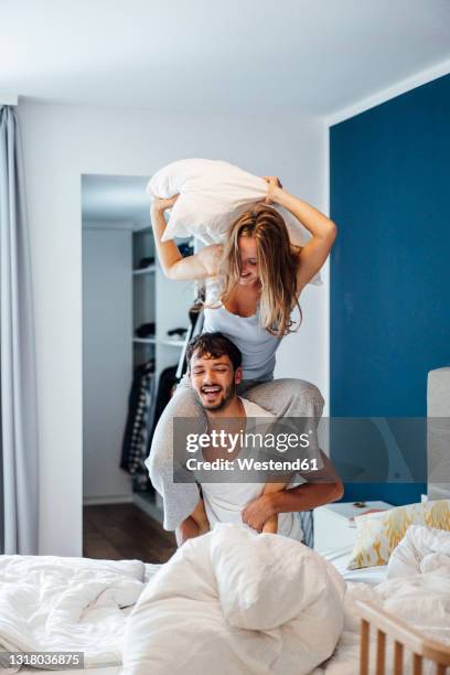 cheerful young woman having pillow fight with boyfriend in bedroom - kissenschlacht paar stock-fotos und bilder