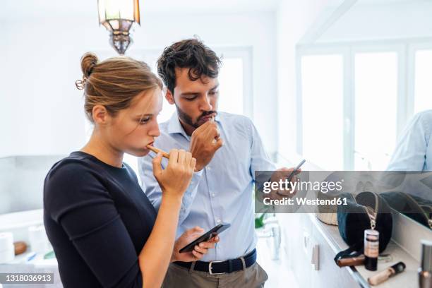 young couple using smart phone while brushing teeth in bathroom - brush teeth phone stock pictures, royalty-free photos & images