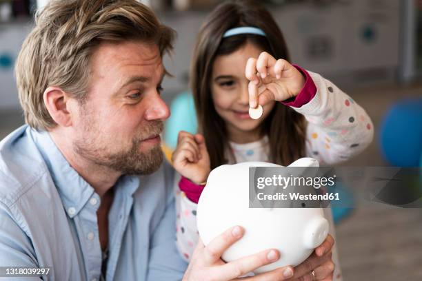 daughter putting coin in piggy bank held by father at home - kids money fotografías e imágenes de stock