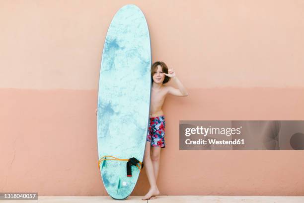 boy wearing swimming trunks gesturing while standing with surfboard in front of beige wall - shaka stock pictures, royalty-free photos & images