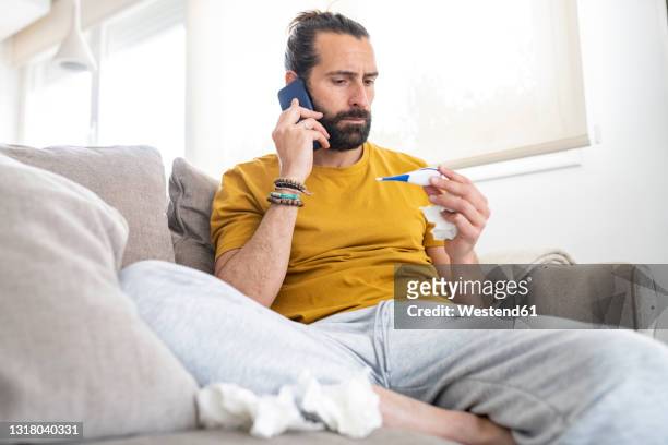 worried man talking on smart phone while looking at thermometer at home - illness stock-fotos und bilder